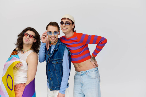 веселі привабливі геї з модними сонцезахисними окулярами, що тримають веселковий прапор і посміхаються на камеру - Фото, зображення