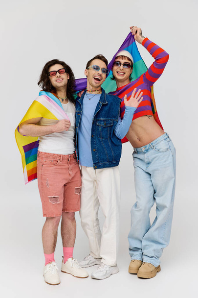 joyful appealing gay men with trendy sunglasses holding rainbow flag and smiling at camera - Фото, изображение