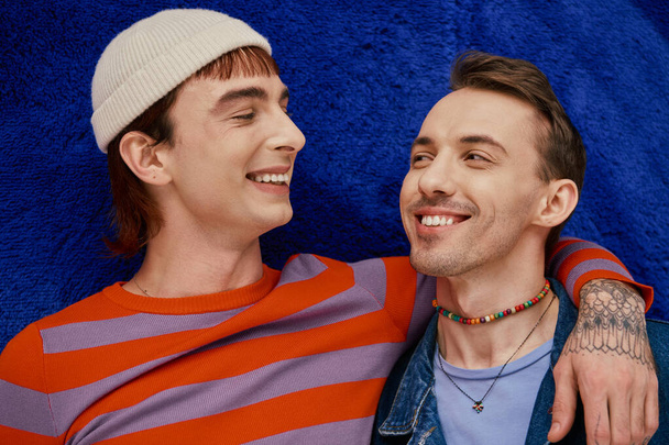 two cheerful good looking gay men in vibrant clothes posing on dark blue backdrop, pride month - Photo, Image