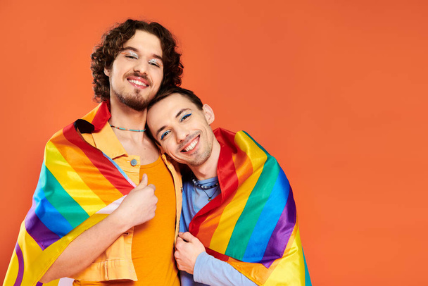 zadowolony atrakcyjny młody gej mężczyźni w przytulne stroje z tęczową flagą na pomarańczowym tle, duma miesiąc - Zdjęcie, obraz