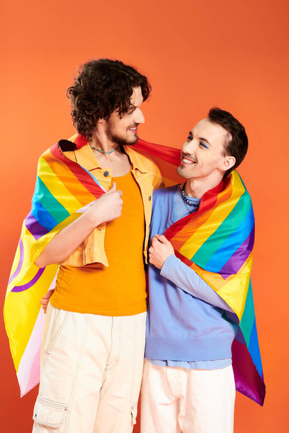 радісні привабливі молоді геї в затишних вбраннях з веселковим прапором на помаранчевому тлі, місяць гордості - Фото, зображення