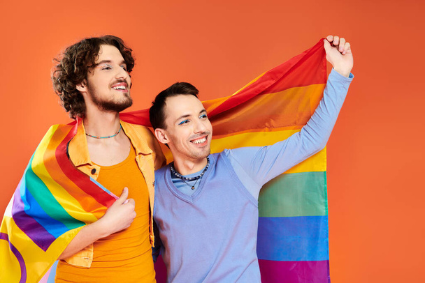 wesoły atrakcyjny młody gej mężczyźni w przytulne stroje z tęczową flagą na pomarańczowym tle, duma miesiąc - Zdjęcie, obraz