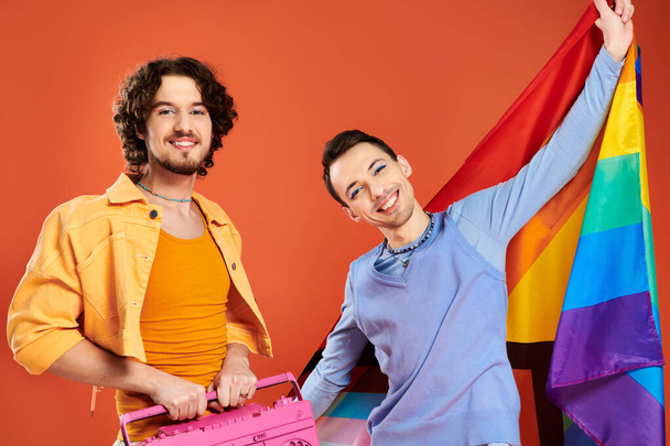 dos contento guapo gay amigos posando con cinta grabadora y arco iris bandera en naranja telón de fondo - Foto, Imagen