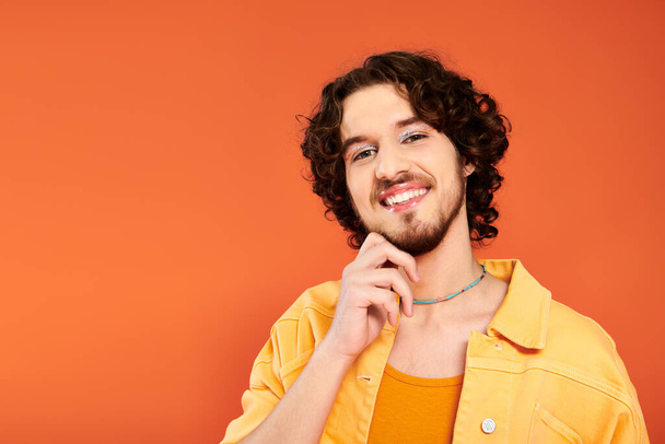 jolly appealing gay man with dark hair and vibrant makeup posing on orange backdrop, pride month - Foto, afbeelding