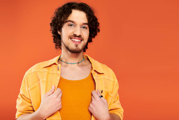 merry appealing gay man with dark hair and vibrant makeup posing on orange backdrop, pride month - Photo, Image