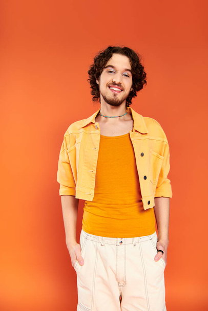 positive appealing gay man with dark hair and vibrant makeup posing on orange backdrop, pride month - Zdjęcie, obraz