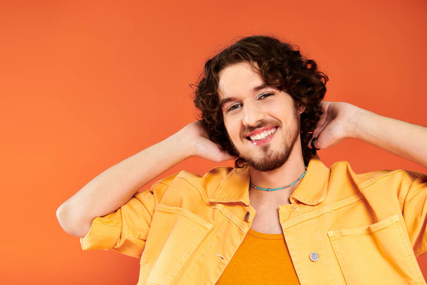 joyous appealing gay man with dark hair and vibrant makeup posing on orange backdrop, pride month - Фото, изображение