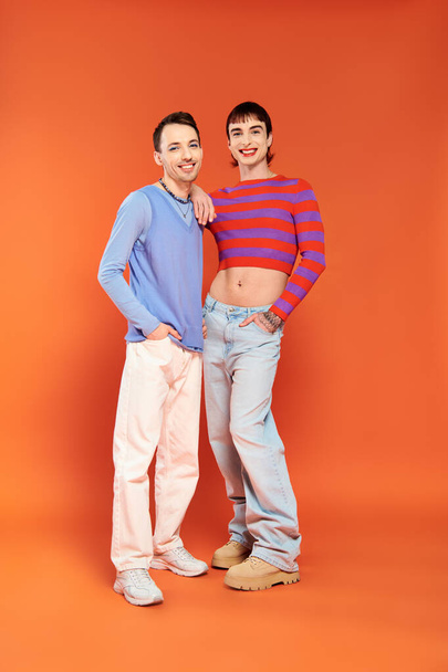 two cheerful young gay men with vibrant makeup in stylish attires posing and smiling at camera - Photo, Image