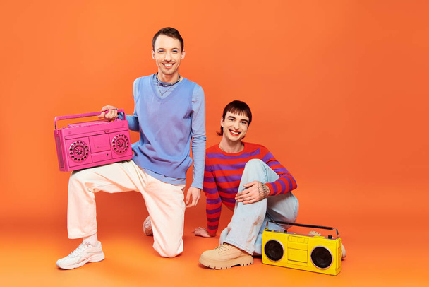 two cheerful good looking gay men with vibrant makeup posing with tape recorders on orange backdrop - Photo, Image