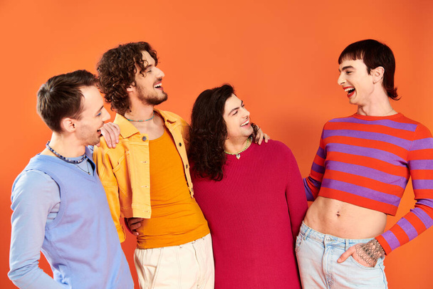 τέσσερις ελκυστικούς χαρούμενους γκέι άνδρες με ζωντανά ρούχα ποζάρουν μαζί σε πορτοκαλί φόντο, μήνα υπερηφάνειας - Φωτογραφία, εικόνα