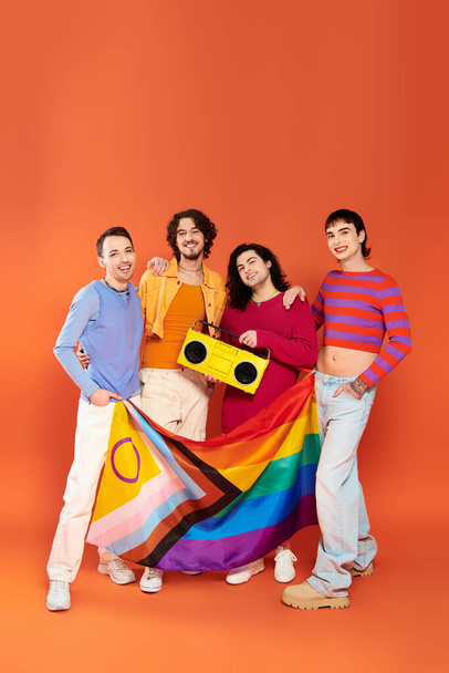 four young merry gay friends posing with rainbow flag and tape recorder on orange background - Photo, Image