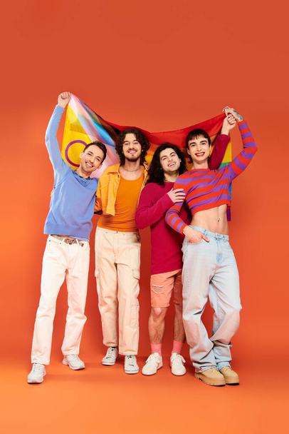 カメラの前に虹の旗を掲げている活気に満ちた服装の4人の楽しいファッショナブルなゲイ男性,誇り - 写真・画像