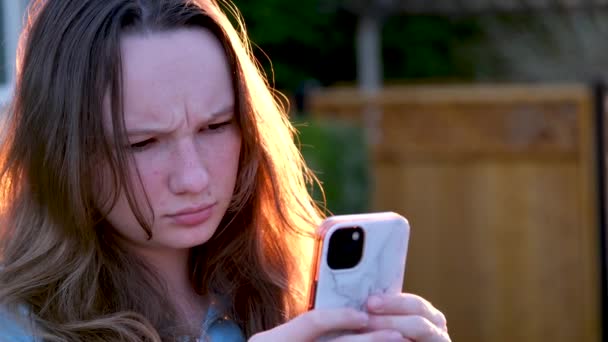una adolescente está insatisfecha con algo que frunció el ceño sus cejas en la puesta de sol de la calle una chica mira el teléfono un nuevo mensaje información desagradable  - Metraje, vídeo