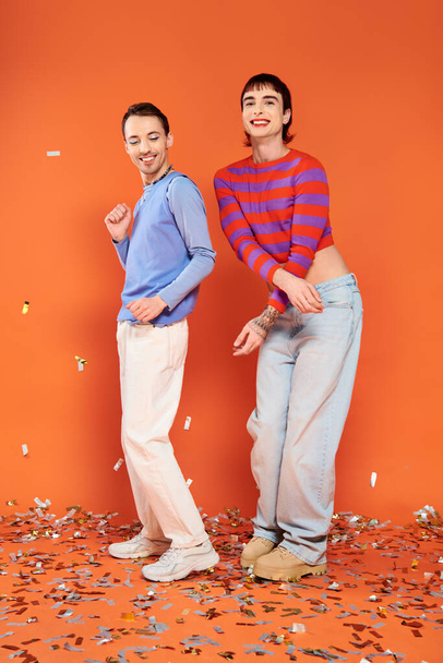 two cheerful stylish gay men in vibrant attires having fun under confetti rain on orange background - Photo, Image