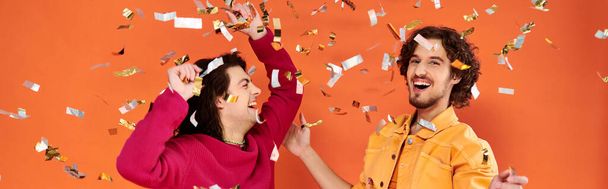 joyeux élégant gay les hommes dans vibrant attires avoir amusant sous confettis pluie sur orange toile de fond, bannière - Photo, image