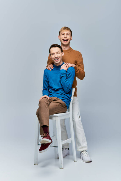 Δύο άντρες, ένα ερωτευμένο γκέι ζευγάρι με περιστασιακή ενδυμασία, κάθονται σε ένα σκαμνί ποζάροντας για την κάμερα σε ένα γκρι φόντο.. - Φωτογραφία, εικόνα