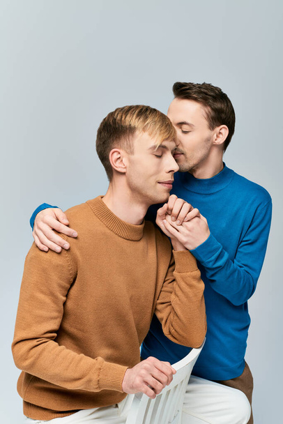 Dva muži, milující homosexuální pár, sedící spolu na židli v nenucených šatech na šedém pozadí. - Fotografie, Obrázek