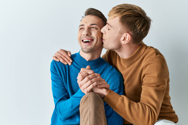 A gay couple in casual attire sitting closely together on a gray backdrop. - Photo, Image