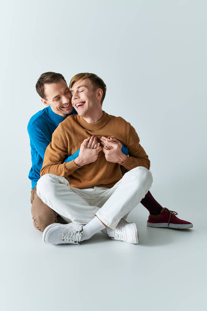 Dos hombres con atuendo casual, sentados en el suelo, compartiendo un abrazo. - Foto, Imagen