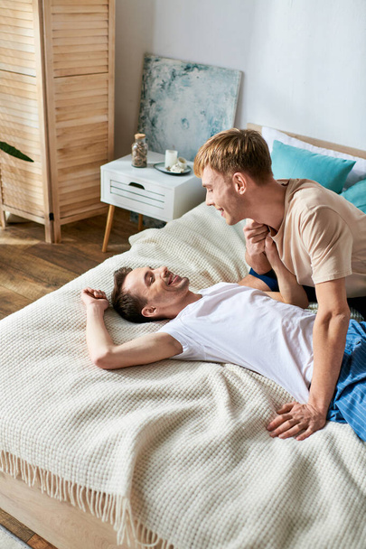 Δύο άνδρες με περιστασιακή ενδυμασία μοιράζονται μια οικεία στιγμή σε ένα κρεβάτι. - Φωτογραφία, εικόνα