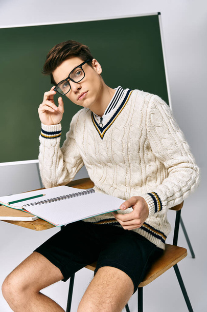 Ein junger Mann mit Brille sitzt an einem Schreibtisch mit einem Notizbuch, tief in Gedanken. - Foto, Bild