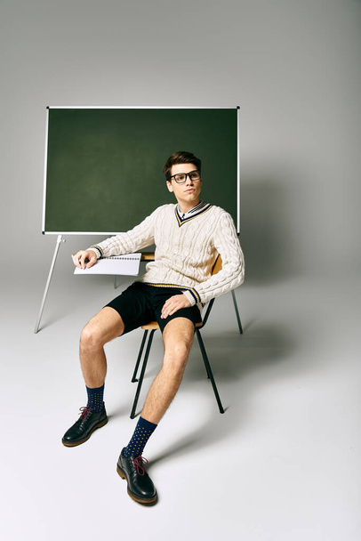Un bellissimo studente in uniforme seduto su una sedia accanto a una lavagna verde. - Foto, immagini