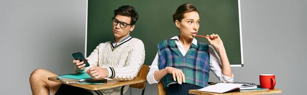 Zwei Studenten, ein Mann und eine Frau, sitzen an einem Schreibtisch vor einer Tafel in einem College-Umfeld. - Foto, Bild