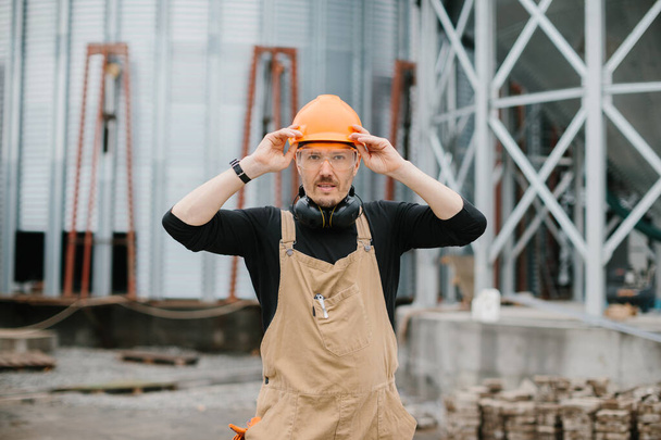 Будівельник в захисному шоломі, окулярах і спецодязі стоїть на тлі будівництва зернового силосу. - Фото, зображення
