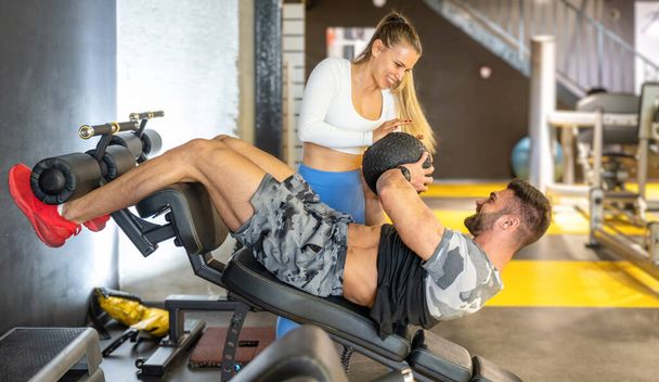 Młoda kobieta trener osobisty pomaga muskularny mężczyzna podczas wykonywania chrupania z piłką fitness na ławce treningowej na siłowni. - Zdjęcie, obraz