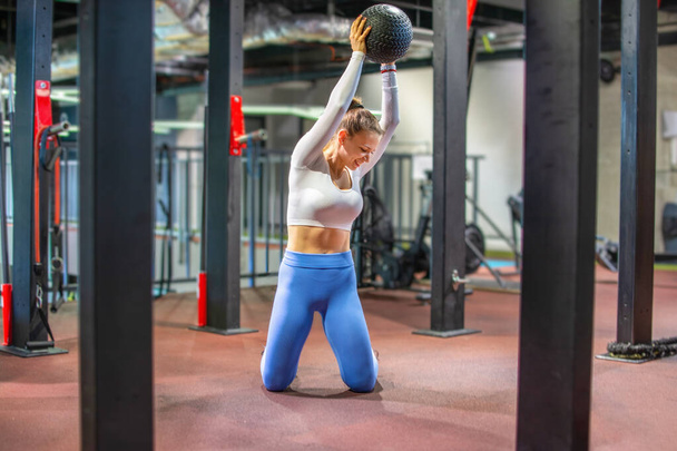 Привлекательная молодая женщина, стоящая на коленях и занимающаяся спортом с мячом в спортзале. - Фото, изображение