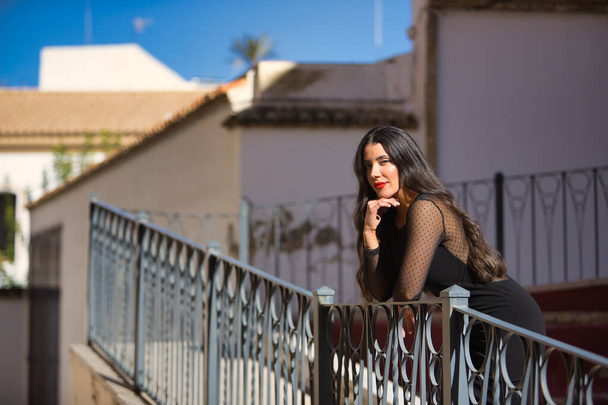 Mujer joven, guapa, morena en un elegante vestido negro, posando mirando a la cámara apoyada en una barandilla en un hermoso pueblo blanco andaluz. Concepto belleza, moda, tendencia, viajes, rural. - Foto, imagen
