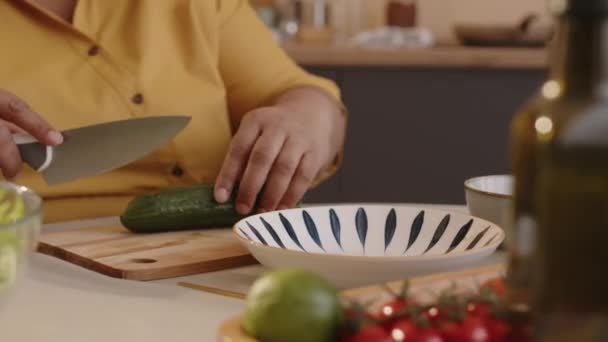 Наклон пожилой черной женщины сидя за столом приготовления пищи и резки свежий огурец на деревянной доске с ножом, делая здоровый овощной салат на обед дома - Кадры, видео