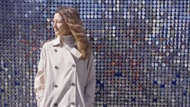 Şık bir trençkot giymiş genç bir kadın düşünceli bir şekilde dikiliyor, yansıtıcı disklerin ışıltılı arka planına karşı duran, kentsel karmaşıklığın bir anını yakalıyor.. - Video, Çekim