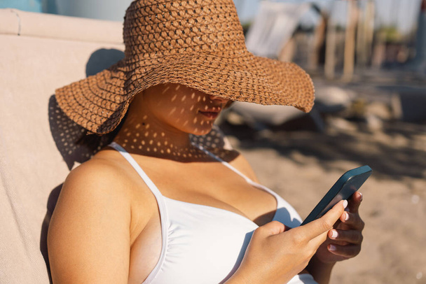 Egy vidám fiatal nő széles karimájú szalmakalapot visel, és egy fehér bikini szelfit készít az okostelefonjával, élvezve a fényes napsütést az óceán partján, tiszta kék égbolttal a feje felett.. - Fotó, kép