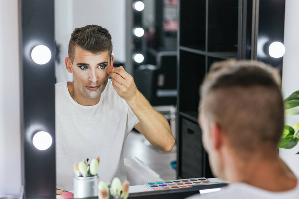 Mężczyzna patrząc w lustro podczas makijażu z gąbką w garderobie. Koncepcja LGBT. - Zdjęcie, obraz
