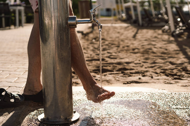 Un uomo si risciacqua i piedi nudi sotto un rubinetto in acciaio inossidabile, le gocce brillano al sole. Un ambiente urbano con una zona sabbiosa che indica la probabile vicinanza a una spiaggia dove le persone possono rinfrescarsi - Foto, immagini