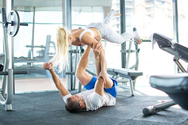 Δυνατός bodybuilder άντρας ξαπλωμένος στην πλάτη του και σηκώνοντας ένα ξανθό κορίτσι γυμναστικής στα πόδια του στο γυμναστήριο - Φωτογραφία, εικόνα