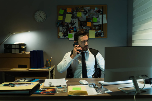 Entschlossener Detektiv bespricht den Fall am Telefon, umgeben von Beweismaterial in einem schwach beleuchteten Büro - Foto, Bild