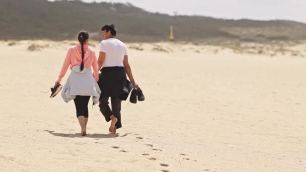 Ένα ζευγάρι που περπατούσε στην παραλία με έναν άντρα που φορούσε λευκό πουκάμισο και μια γυναίκα που φορούσε ροζ μπουφάν. - Πλάνα, βίντεο