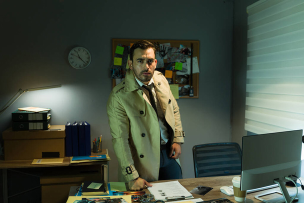 Investigador privado confiante em um casaco de trincheira fica em um escritório mal iluminado, examinando as provas ao seu redor - Foto, Imagem