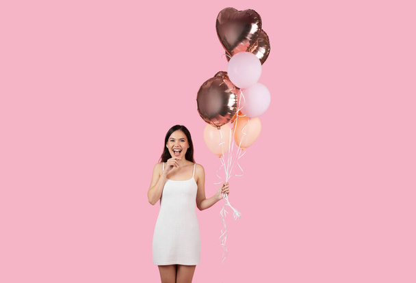 An excited young woman in white dress holds balloons, expression of fun and celebration on a pink background - Photo, Image