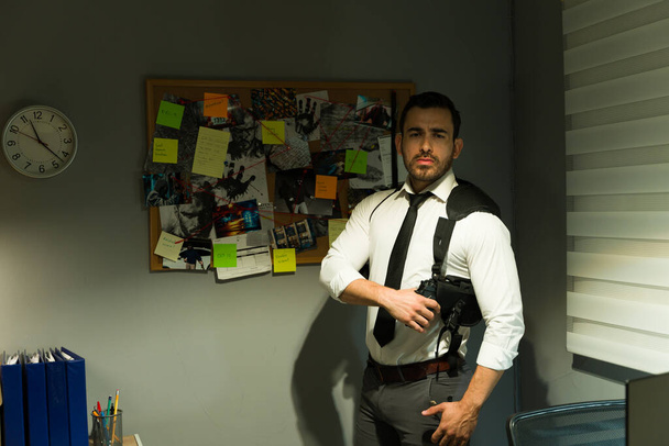 Konzentrierter Ermittler in einem schwach beleuchteten Büro, bewaffnet und vorbereitet neben einer Krimitafel - Foto, Bild