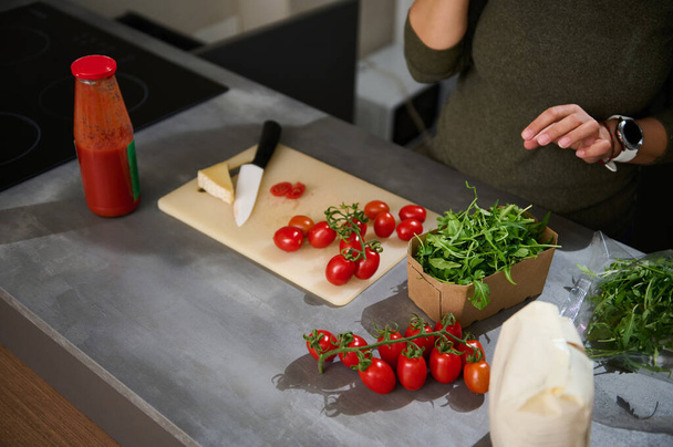 Close-up unknown woman standing at kitchen counter with a fresh bunch of tomato cherry, arugula leaves and Italian pasta in paper packet. Healthy eating, culinary, diet concept. Copy advertising space - Photo, Image
