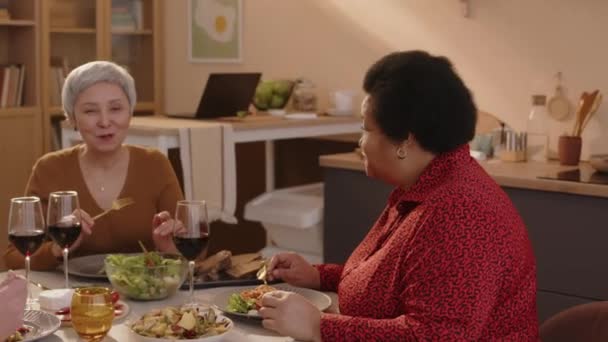 Colpo medio di due donne anziane multietniche e uomo che chiacchierano, bevono vino e cenano insieme in un accogliente appartamento - Filmati, video