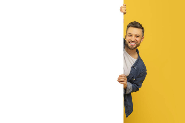 Χαρούμενος γενειοφόρος όμορφος άντρας κρυφοκοιτάζει από το κενό πλακάτ στο κίτρινο φόντο, αντιγράφει την παραλλαγή χώρου - Φωτογραφία, εικόνα
