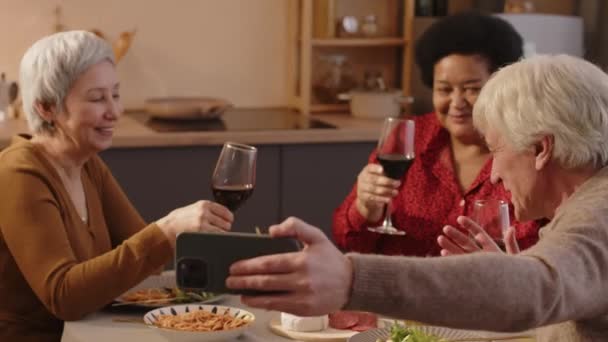 Medium shot of three diverse senior friends laughing and clinking glasses of red wine while trying to take selfie portrait on smartphone sitting at table in bright apartment - Footage, Video