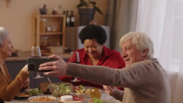Medium shot of group of multiracial senior friends taking selfie portraits on smartphone and having fun, sitting at dinner table together, drinking wine and chatting - Footage, Video