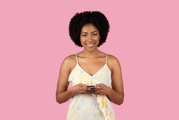 Улыбающаяся молодая афроамериканка с помощью мобильного телефона, одетая в летнее платье на розовом фоне - Фото, изображение