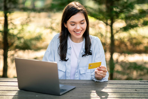 Χαμογελαστή νεαρή γυναίκα που κατέχει πιστωτική κάρτα ενώ χρησιμοποιεί φορητό υπολογιστή, υποδεικνύοντας online αγορές στο δημόσιο πάρκο - Φωτογραφία, εικόνα