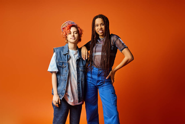 Zwei stilvoll gekleidete junge multikulturelle Freunde, darunter eine nicht binäre Person, stehen selbstbewusst nebeneinander vor einer kühnen orangefarbenen Kulisse. - Foto, Bild